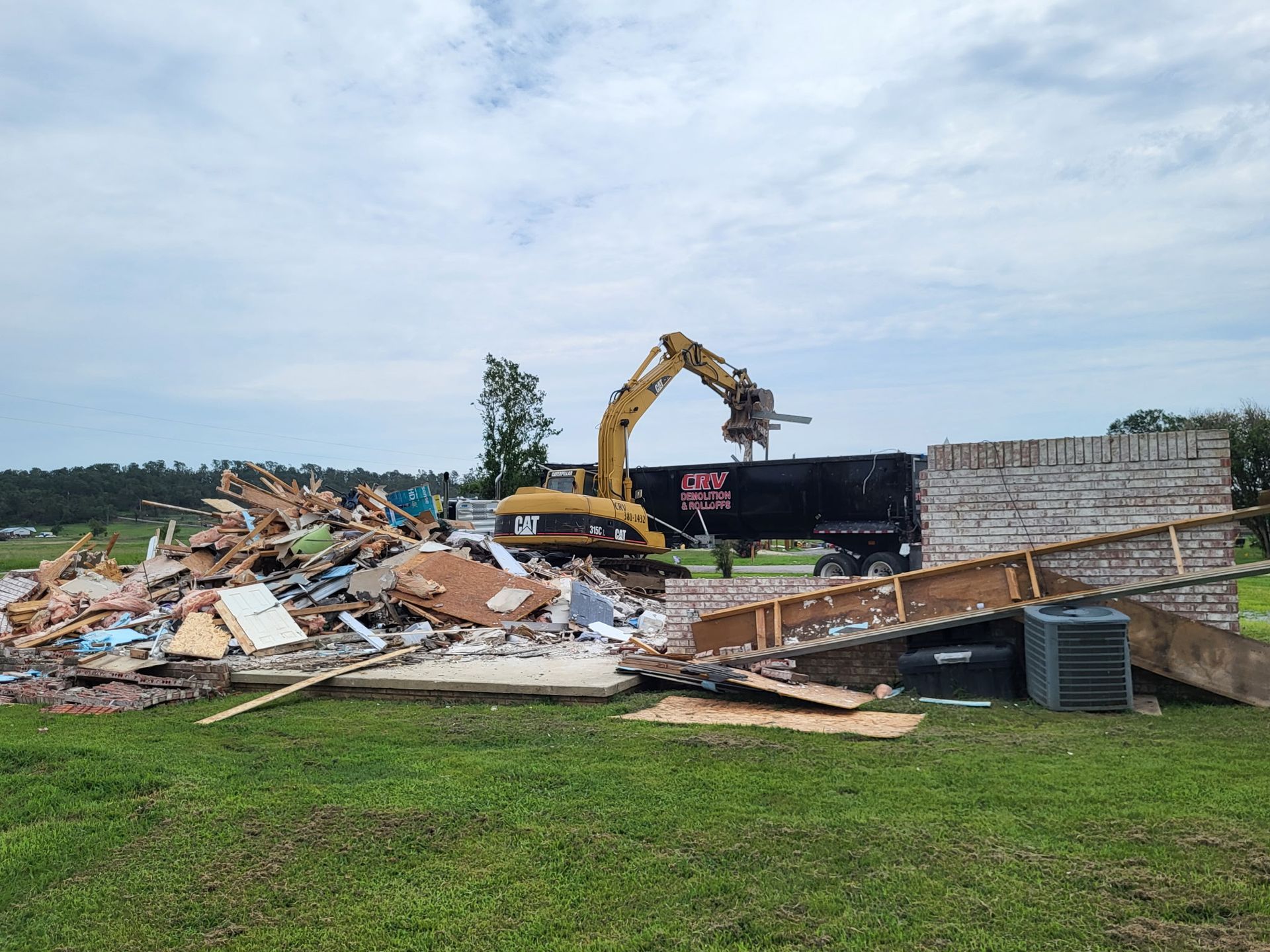 A yellow excavator loads debris from a demolished building into a black dump truck in a grassy field.