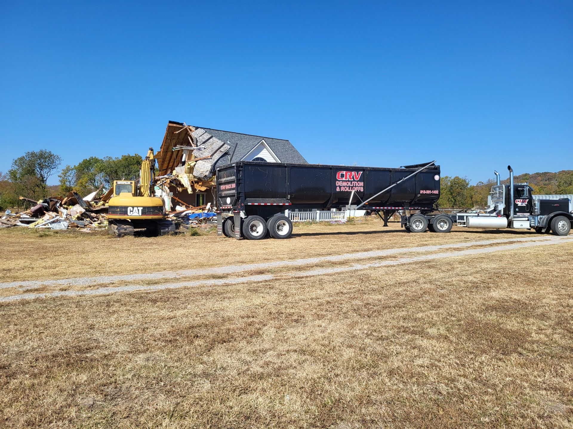 A yellow CAT excavator demoing a house next to a large black dump truck in a field under a clear blue sky.
