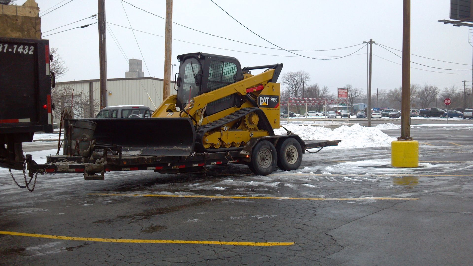 A yellow Caterpillar skid-steer loader sits on a trailer in a snowy, paved parking lot.