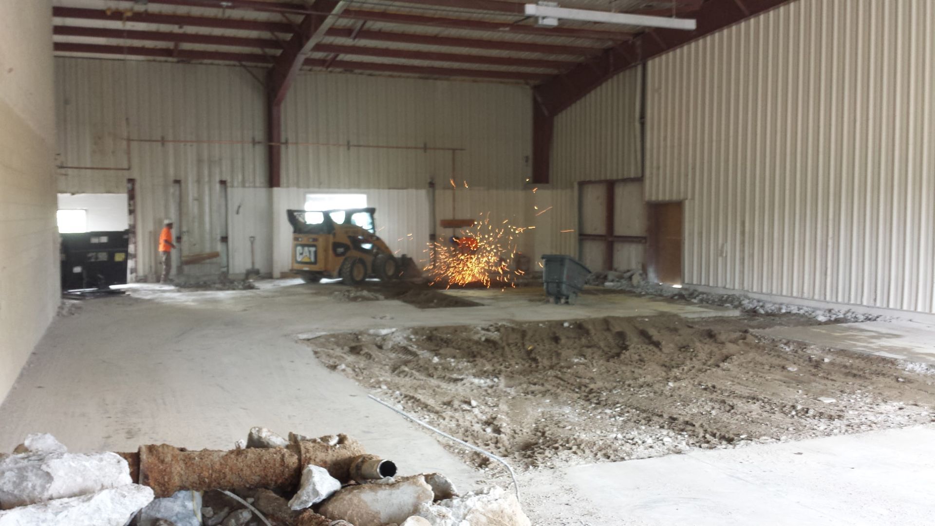 Construction workers using a skid steer to break up concrete in a large, industrial warehouse space.