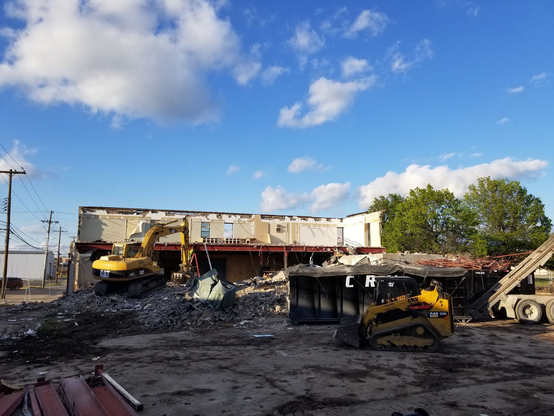 A yellow excavator and a skid steer perform demolition on a large, partially destroyed building under a blue sky.
