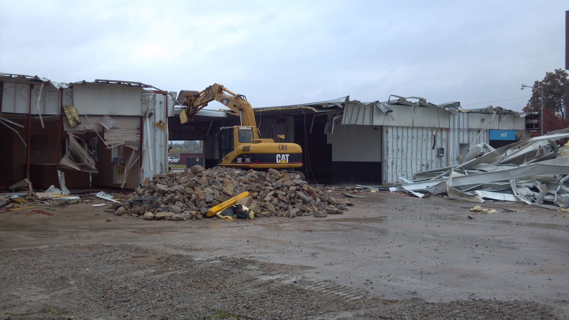 A yellow Caterpillar excavator demolishing a building in a construction zone with a pile of rubble in the foreground.