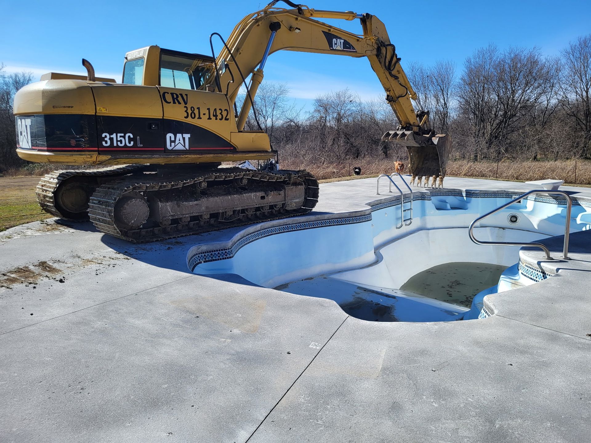 A yellow CAT excavator sits on a concrete deck, positioning its bucket over an empty, white residential swimming pool.