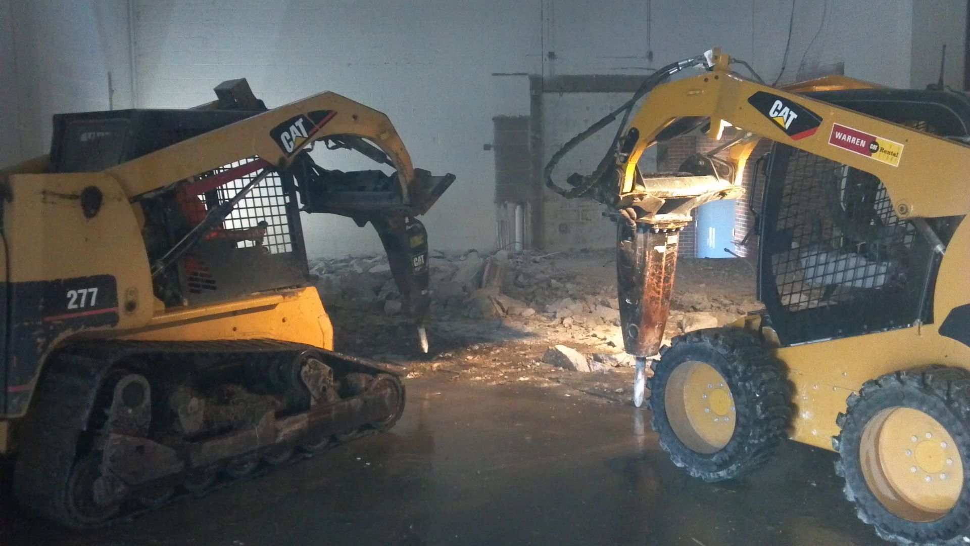 Two yellow Caterpillar skid steer loaders with hydraulic breakers working inside a dimly lit demolition site.