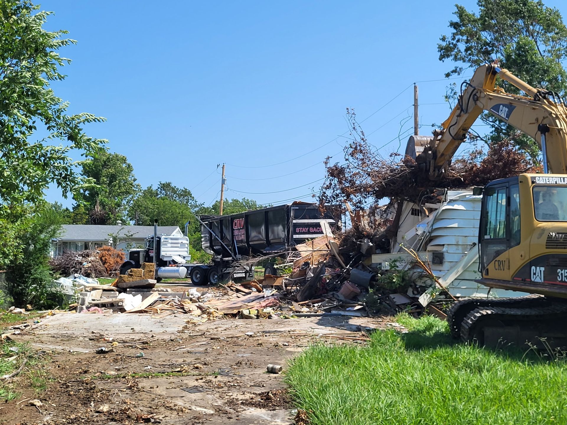 An excavator clears building debris and branches into a large dump truck at a residential demolition site.