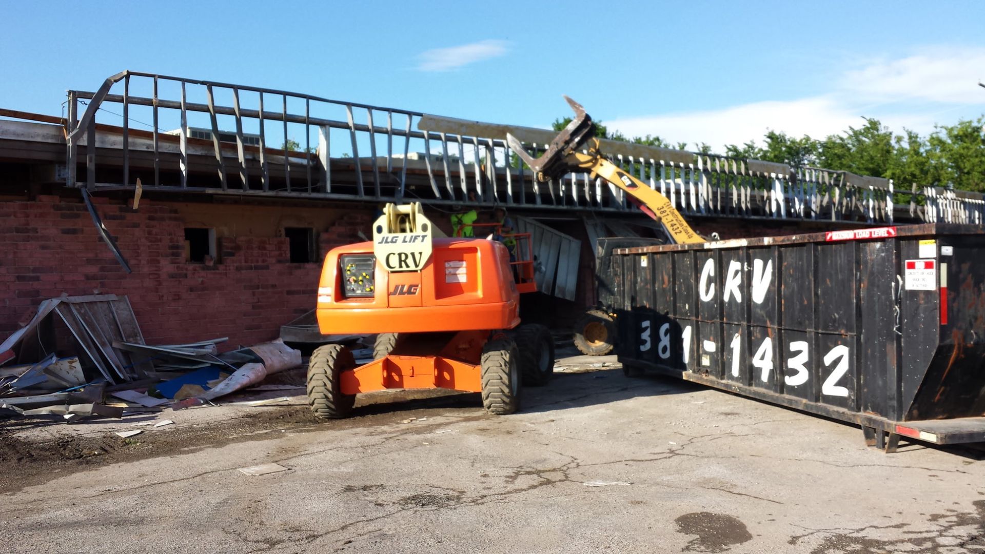 An orange boom lift and a black CRV dumpster are used to demolish a brick building exterior on a sunny day.