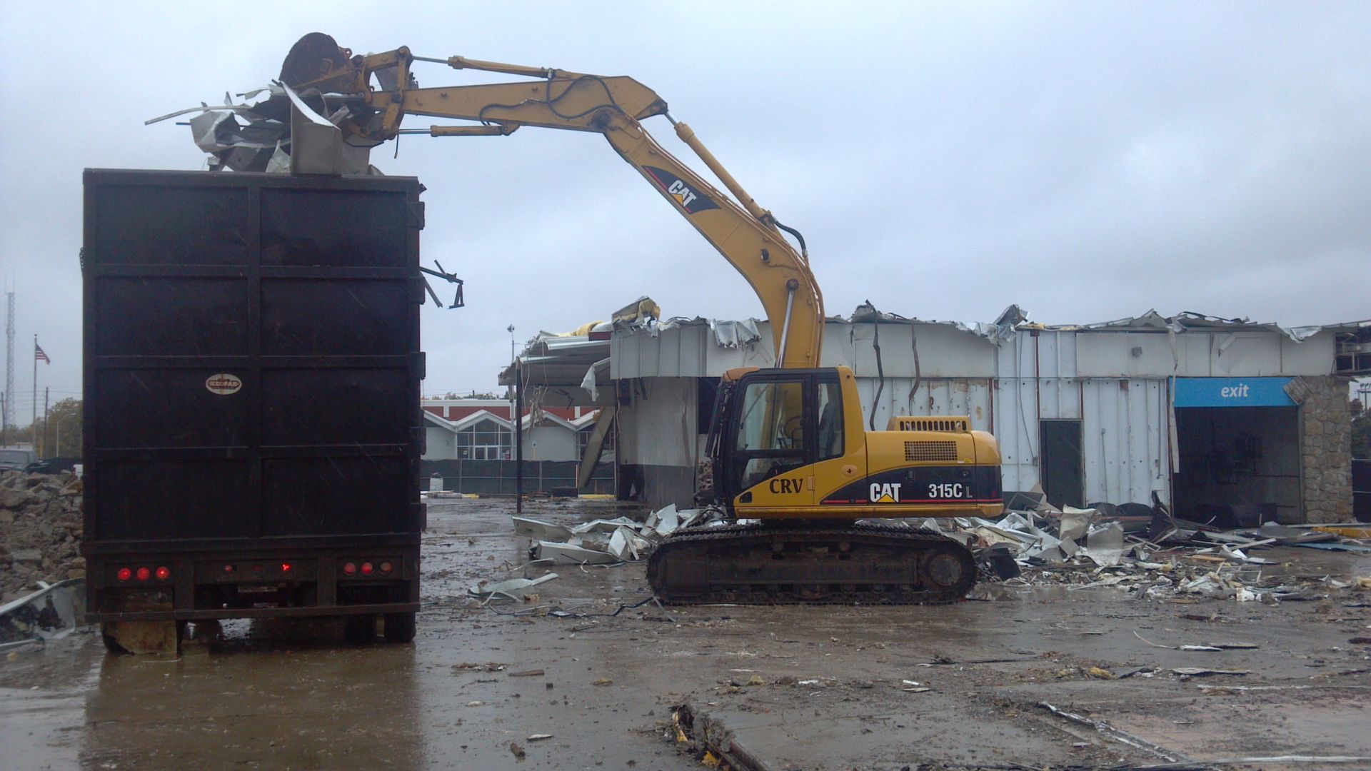 A yellow excavator loads debris into a large black container at a demolition site under a cloudy sky.