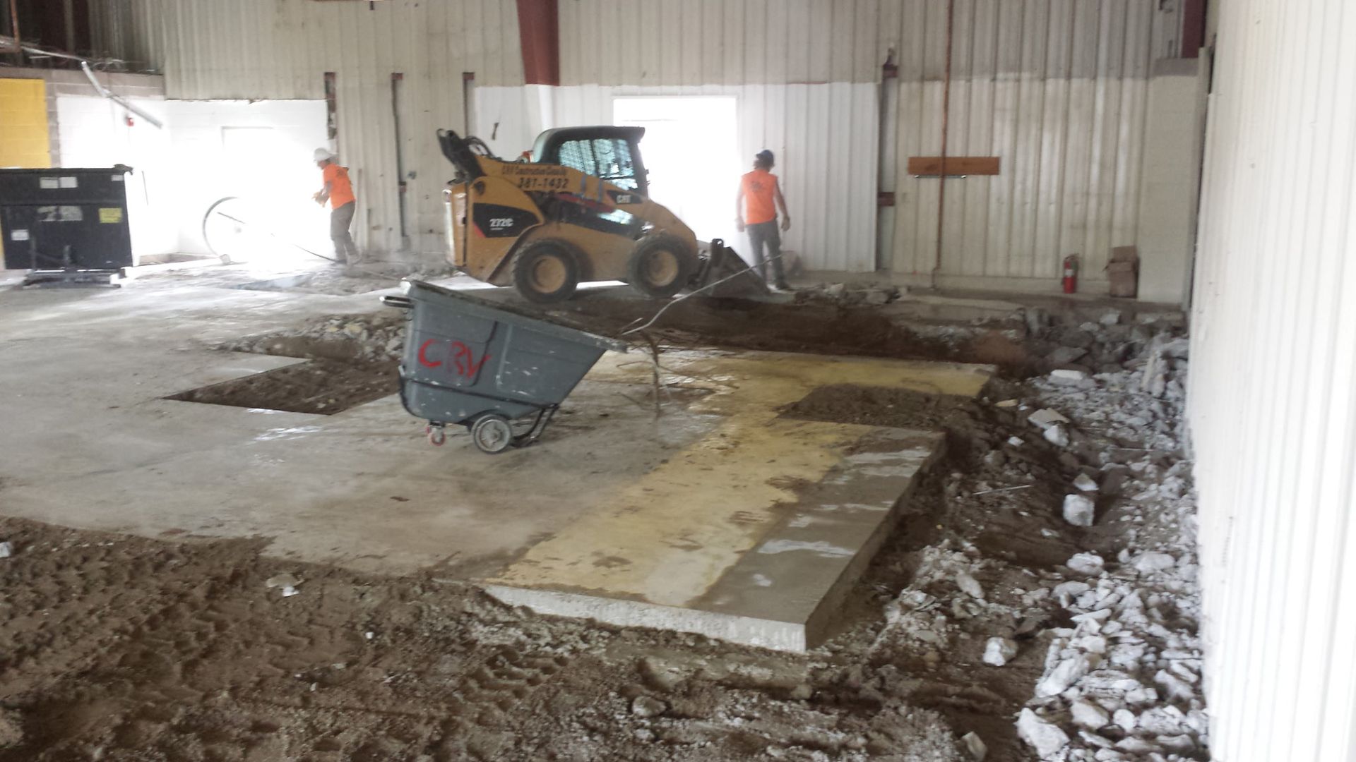 Construction workers operate a skid-steer loader inside a large industrial space with exposed soil and concrete flooring.