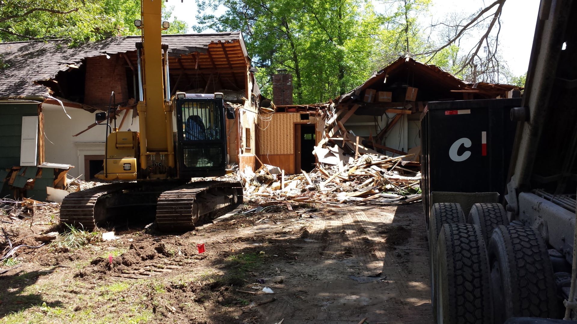 A yellow excavator demolishes a damaged structure, with piles of debris and a large truck visible on a wooded site.