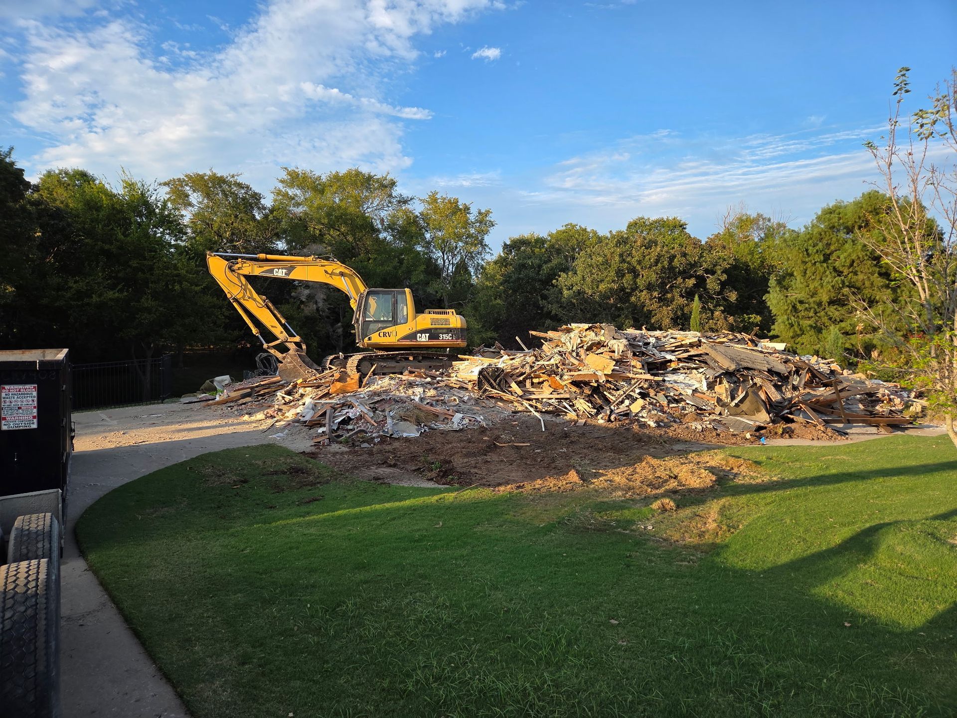 A yellow excavator sits in a grassy yard beside a large pile of rubble from a demolished building.