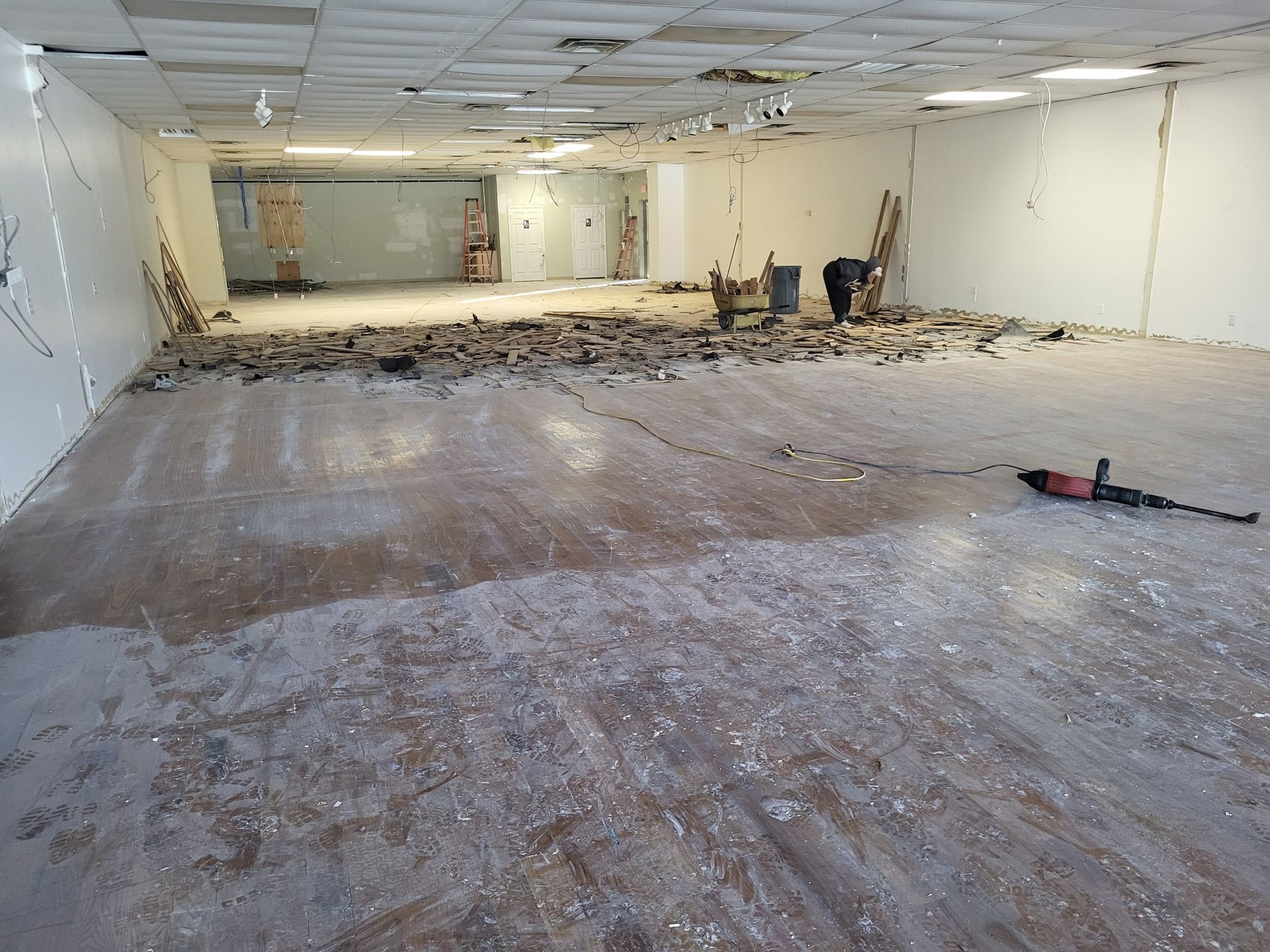 A worker removes debris from a large, unfinished commercial space with exposed flooring and a dropped ceiling.