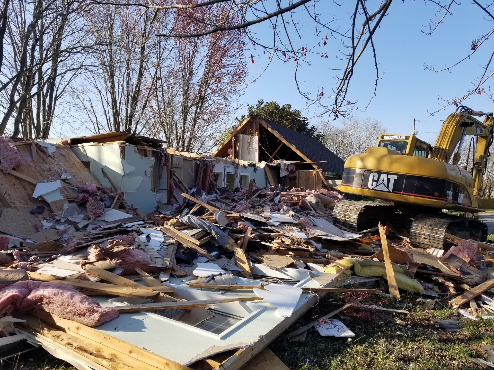 A yellow CAT excavator parked next to a house that is being demolished, leaving a pile of construction debris.