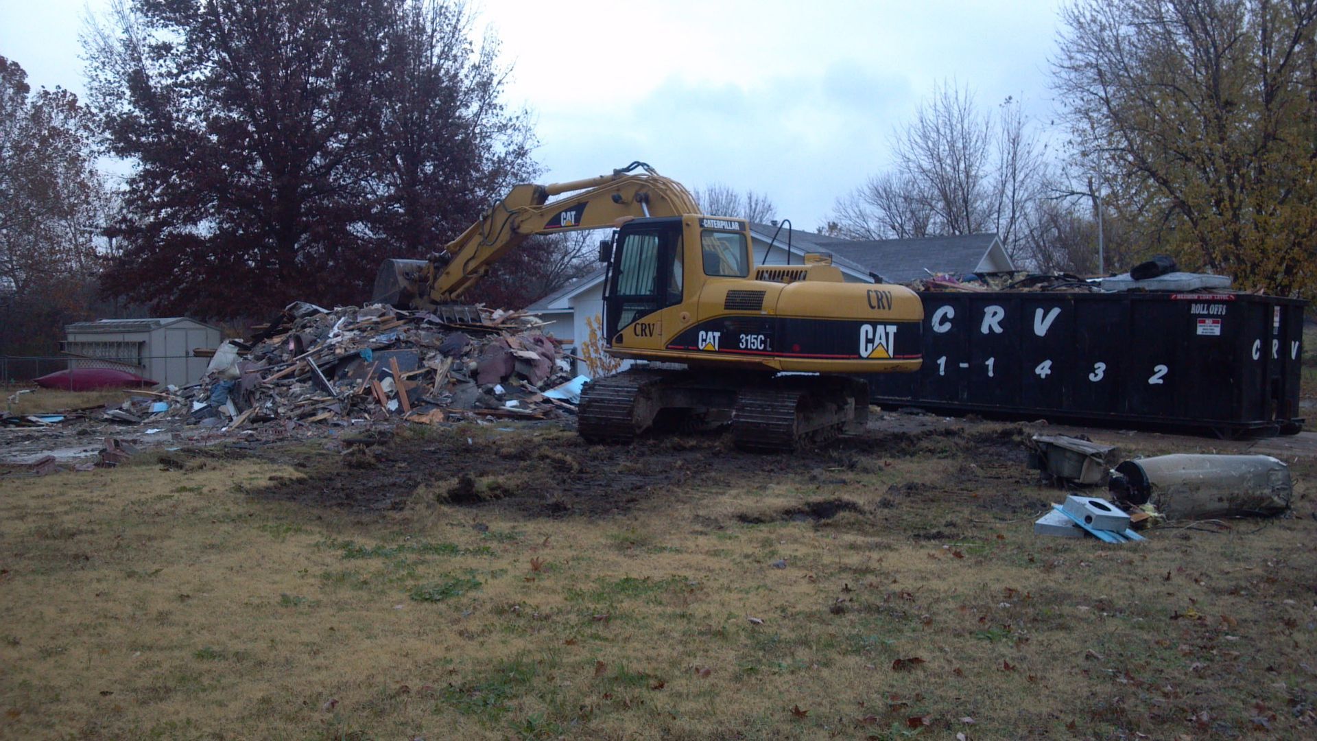 A yellow Caterpillar excavator piles debris next to a large black CRV dumpster in a grassy residential yard.
