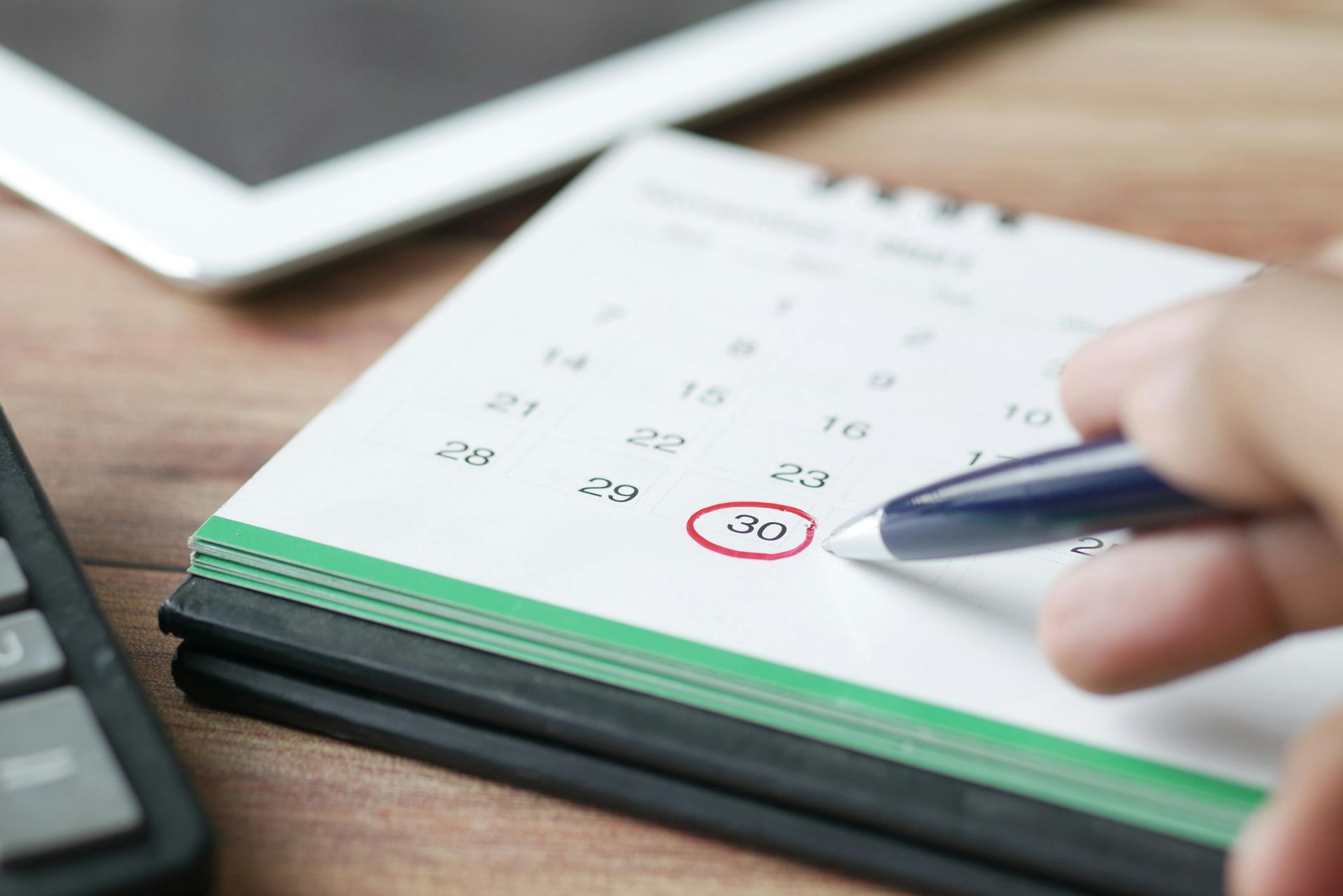 Hand holding a pen over a calendar, circling a date with a laptop in the background