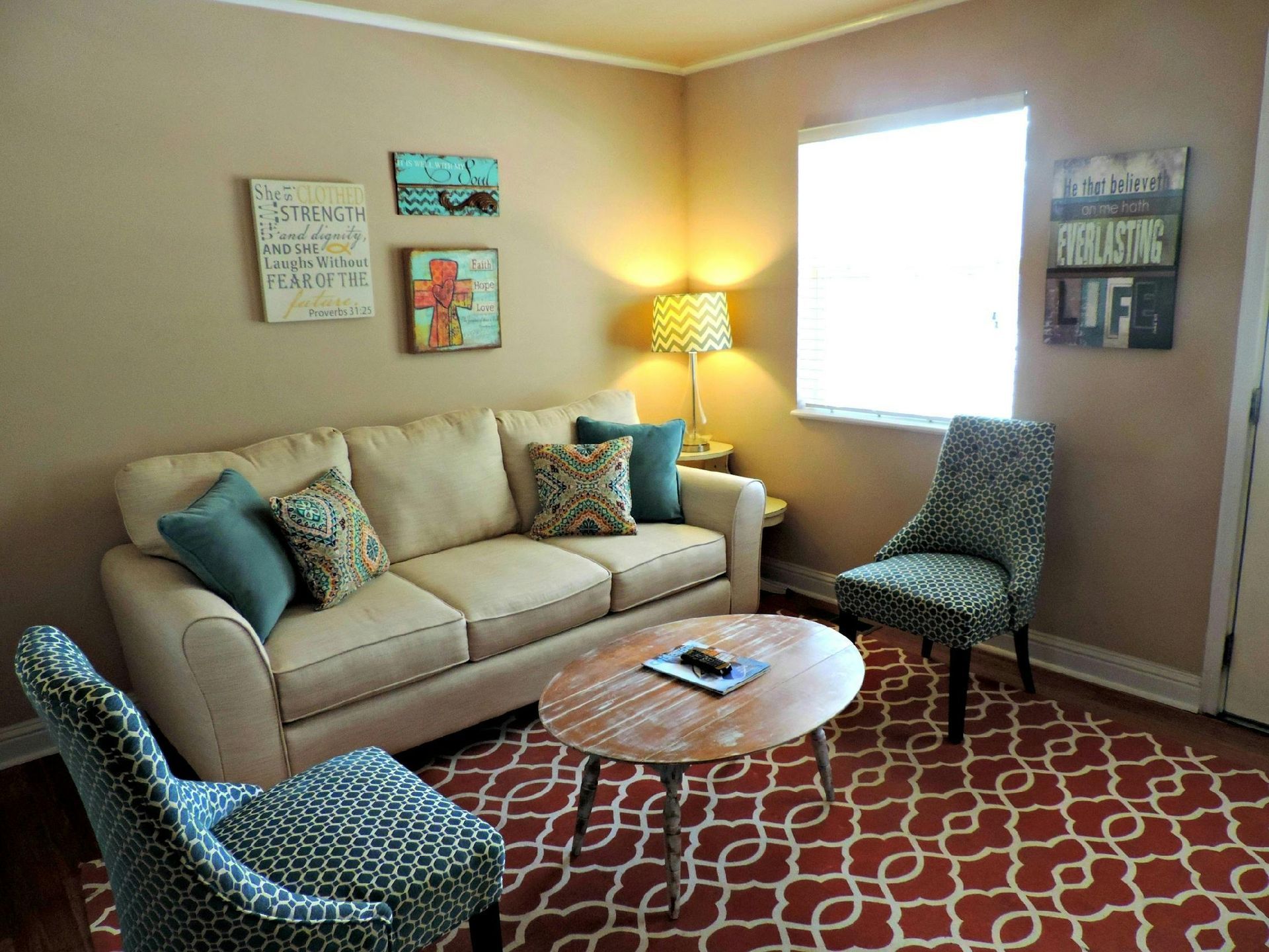 Cozy living room with beige sofa, patterned chairs, round coffee table, red rug, and lamp by a bright window