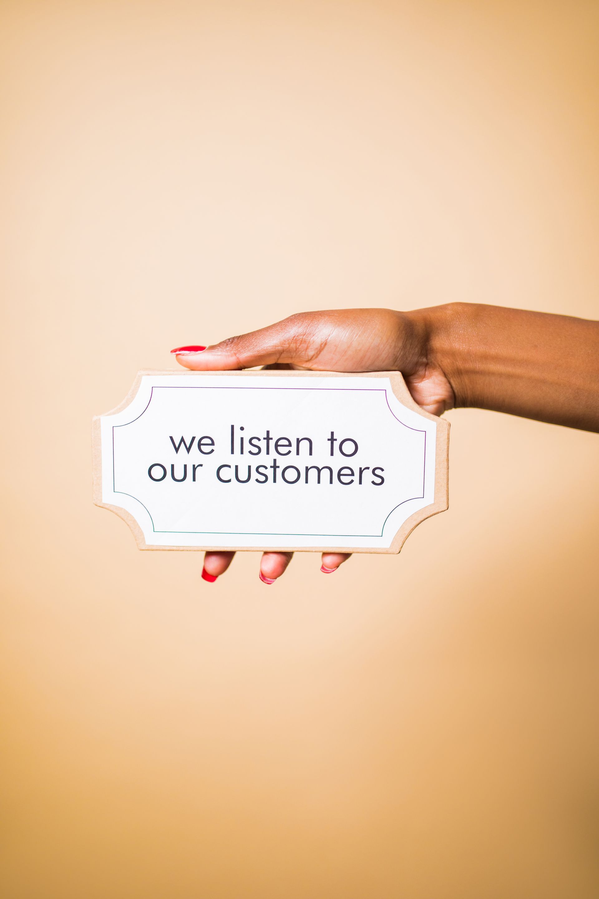 Hand holding a white sign that reads “we listen to our customers” against a beige background