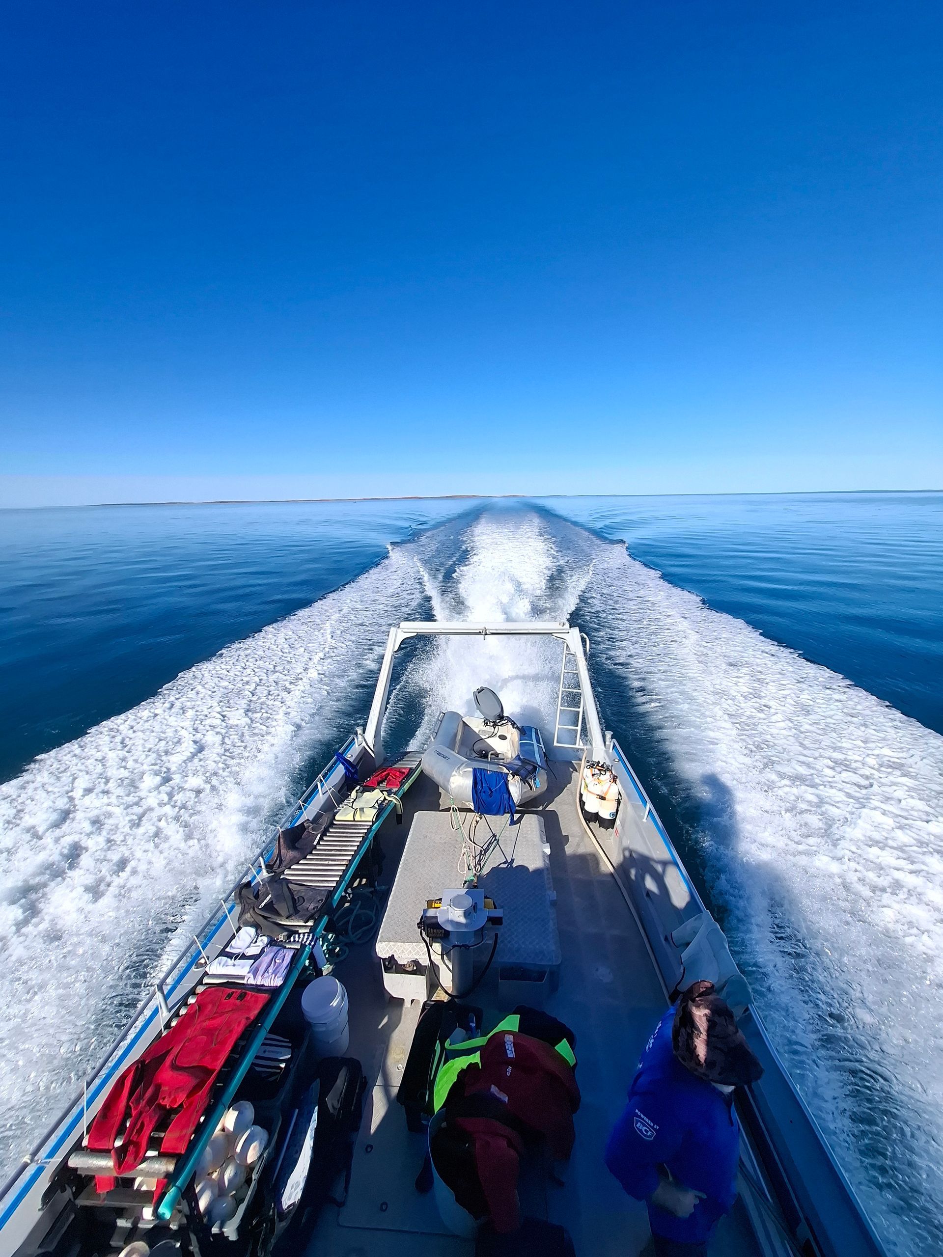 Boat speeding across the ocean, leaving a white wake. Two people in the foreground. Blue sky and water.