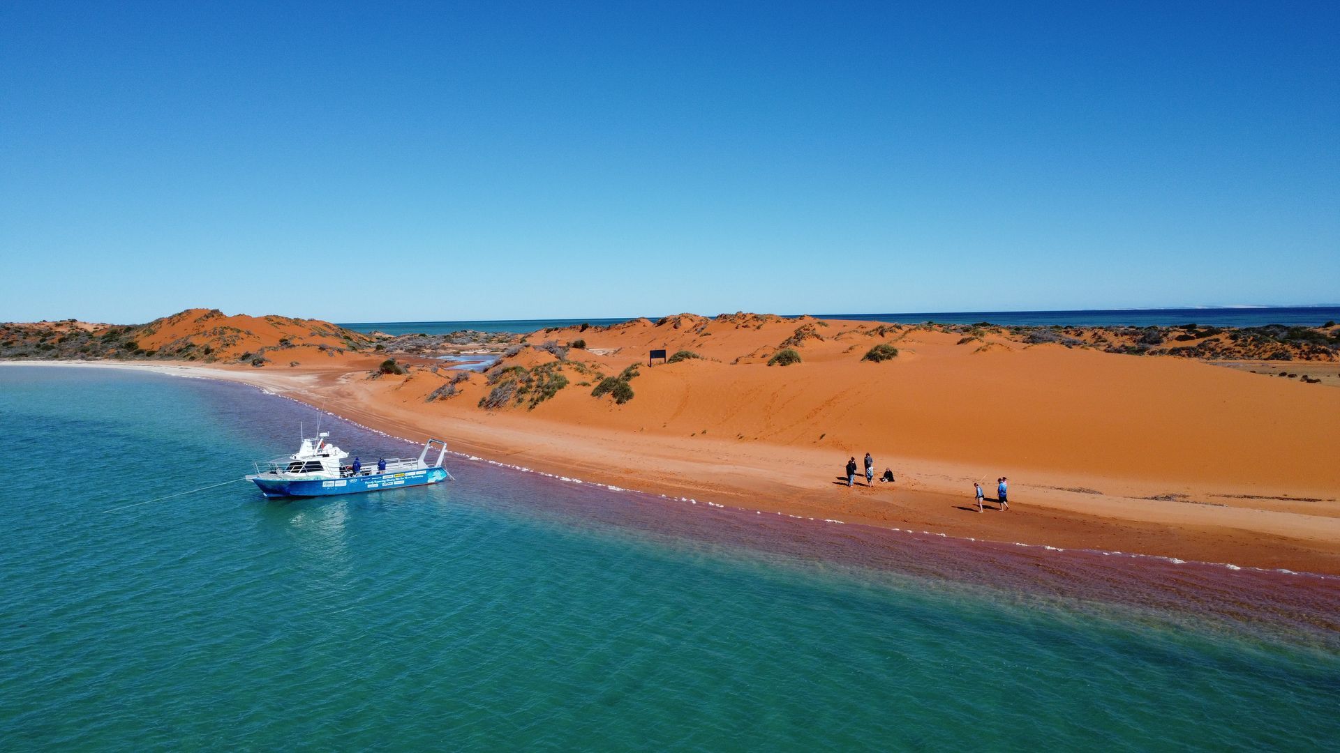 Blue boat with net in turquoise water near a red sand dune beach under a clear blue sky.