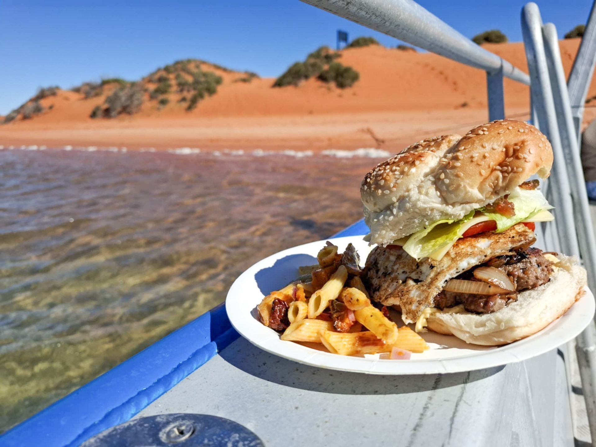 Plate of food: burger and pasta on a boat with red dunes and water in the background.