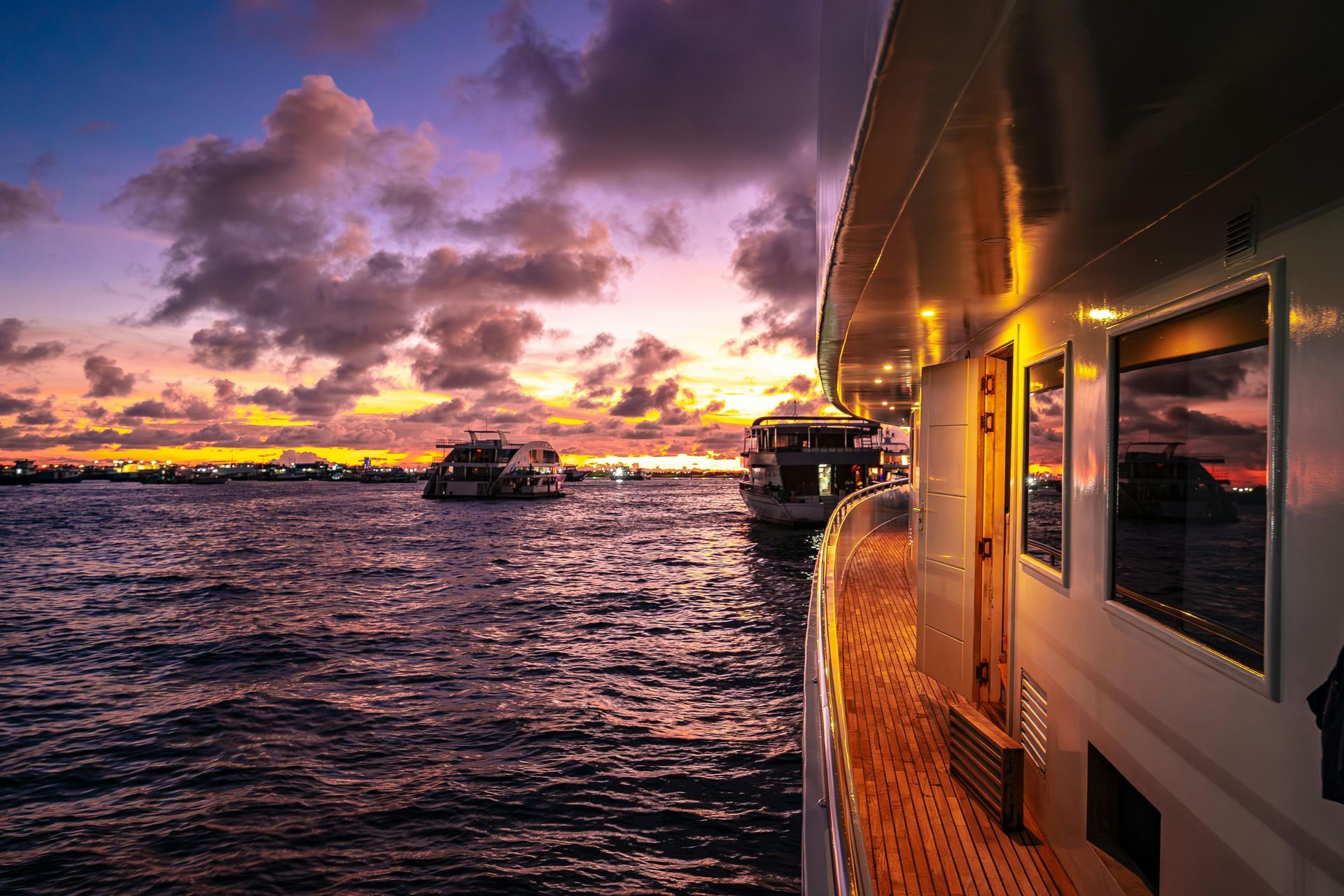 Sunset over water with boats; a yacht's deck and windows in the foreground, colorful sky.