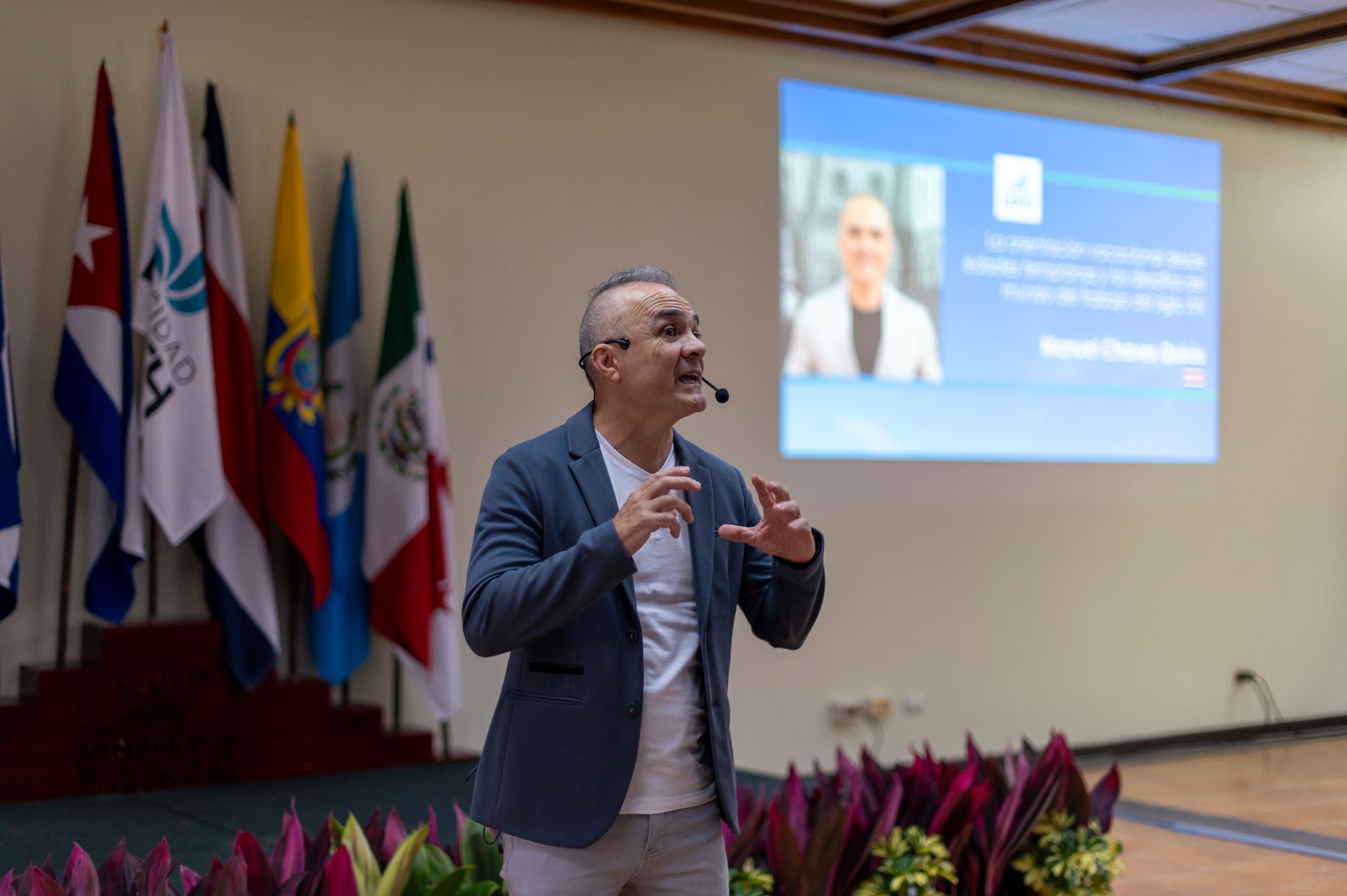 Hombre dando una presentación, gesticulando, con banderas detrás de él y una pantalla que muestra a una mujer.