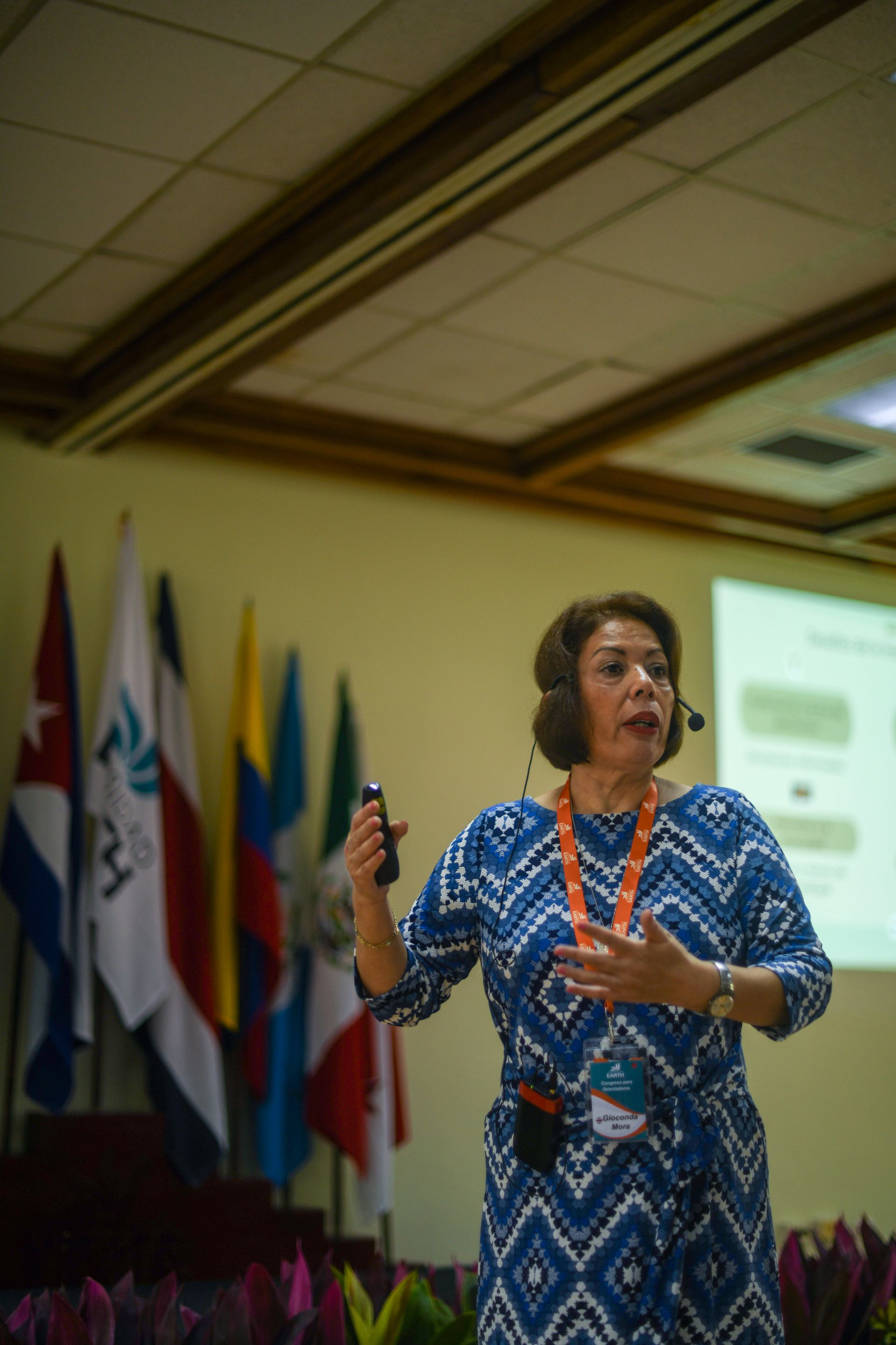 Mujer presentando en una conferencia, gesticulando con un control remoto. Banderas detrás de ella.