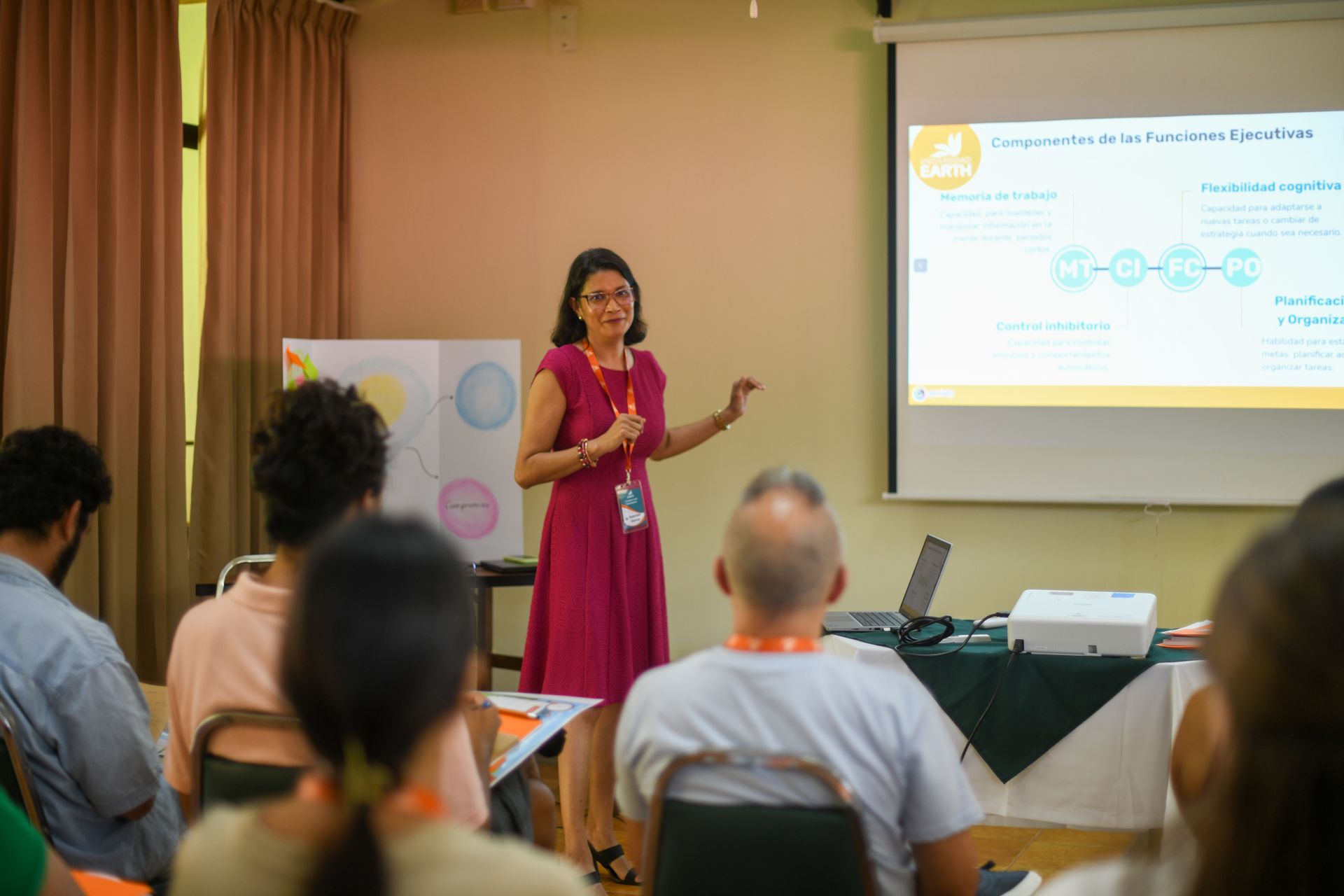 Mujer presentando frente a un grupo; una pantalla de proyector muestra información; la gente está sentada, escuchando.