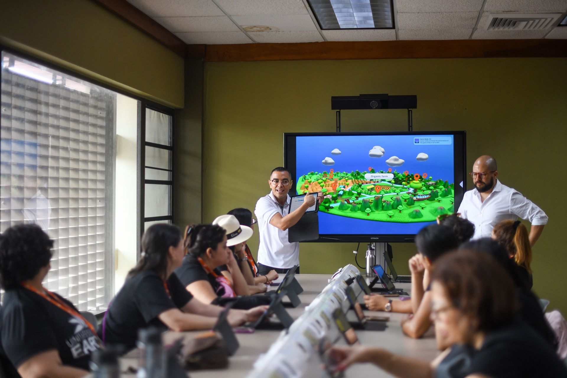 Personas en una sala con una pizarra inteligente, dos hombres están presentando y varios están usando computadoras portátiles.