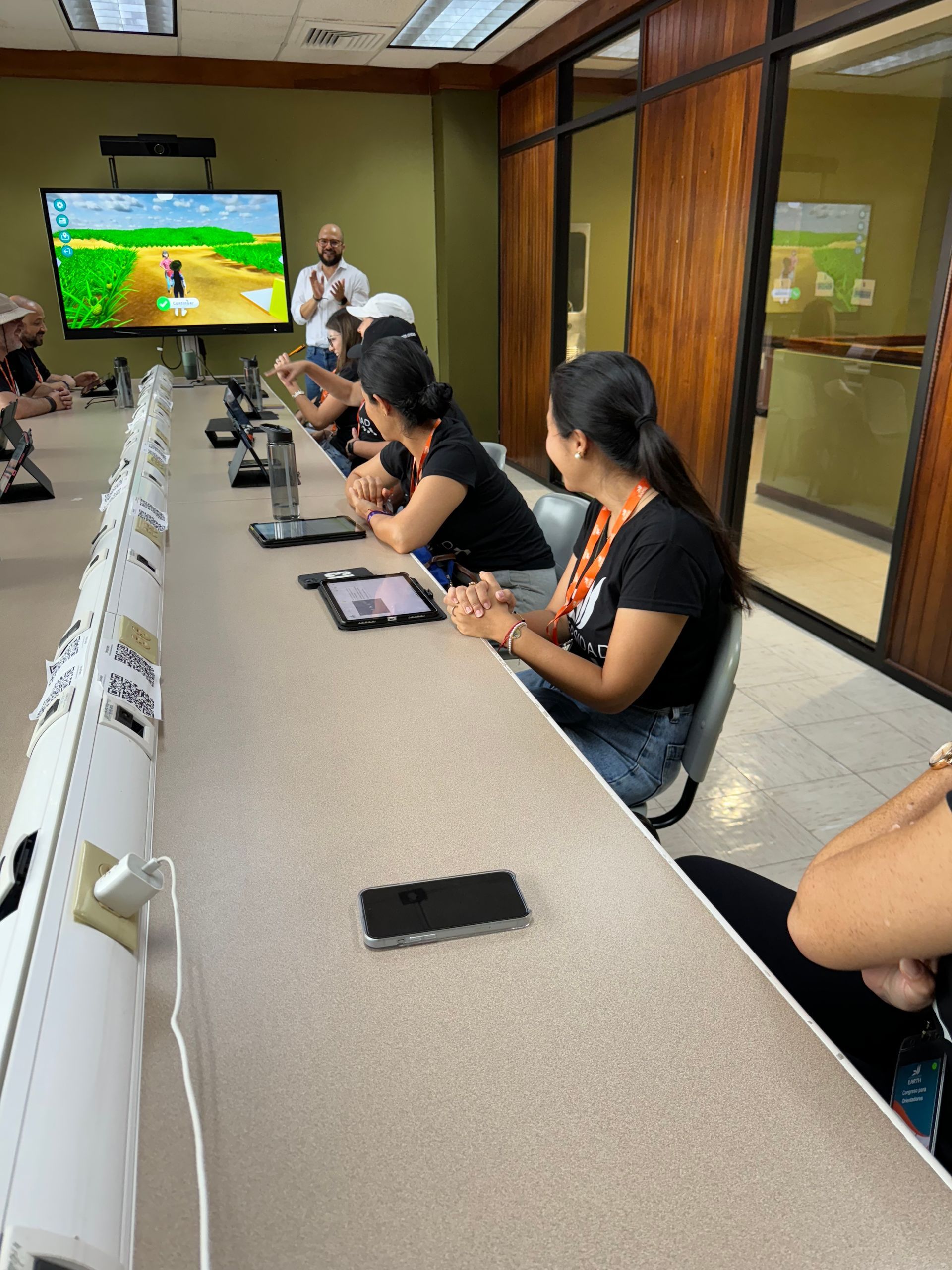 En un aula, las personas ven una presentación en una pantalla. Los estudiantes se sientan en una mesa larga con tabletas.