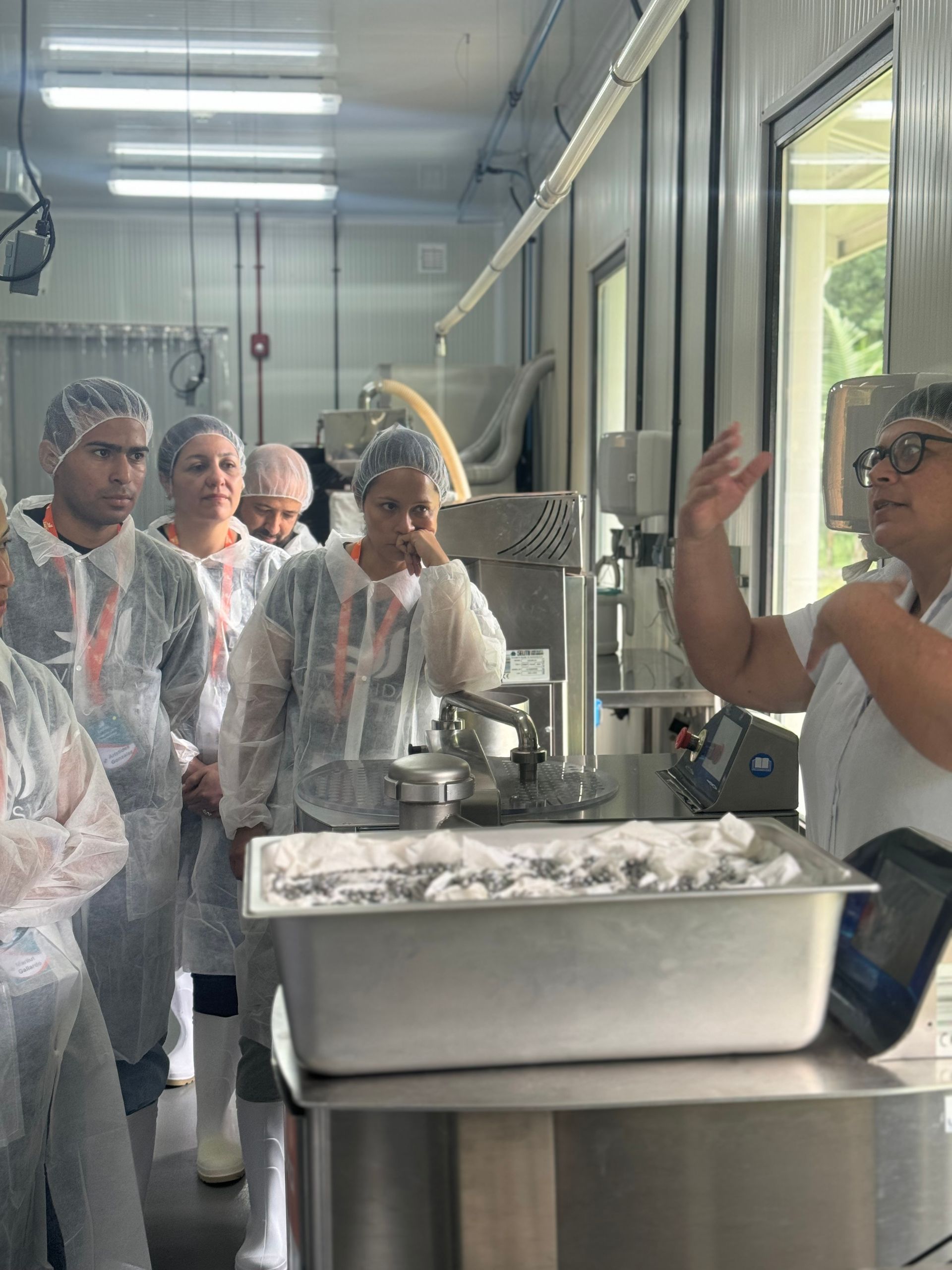 Visita en grupo a una planta de procesamiento de alimentos, observando la maquinaria; una persona gesticula, los demás escuchan.