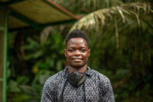 Un hombre de piel oscura sonríe, con una camisa estampada y una mascarilla alrededor del cuello. Fondo verde.