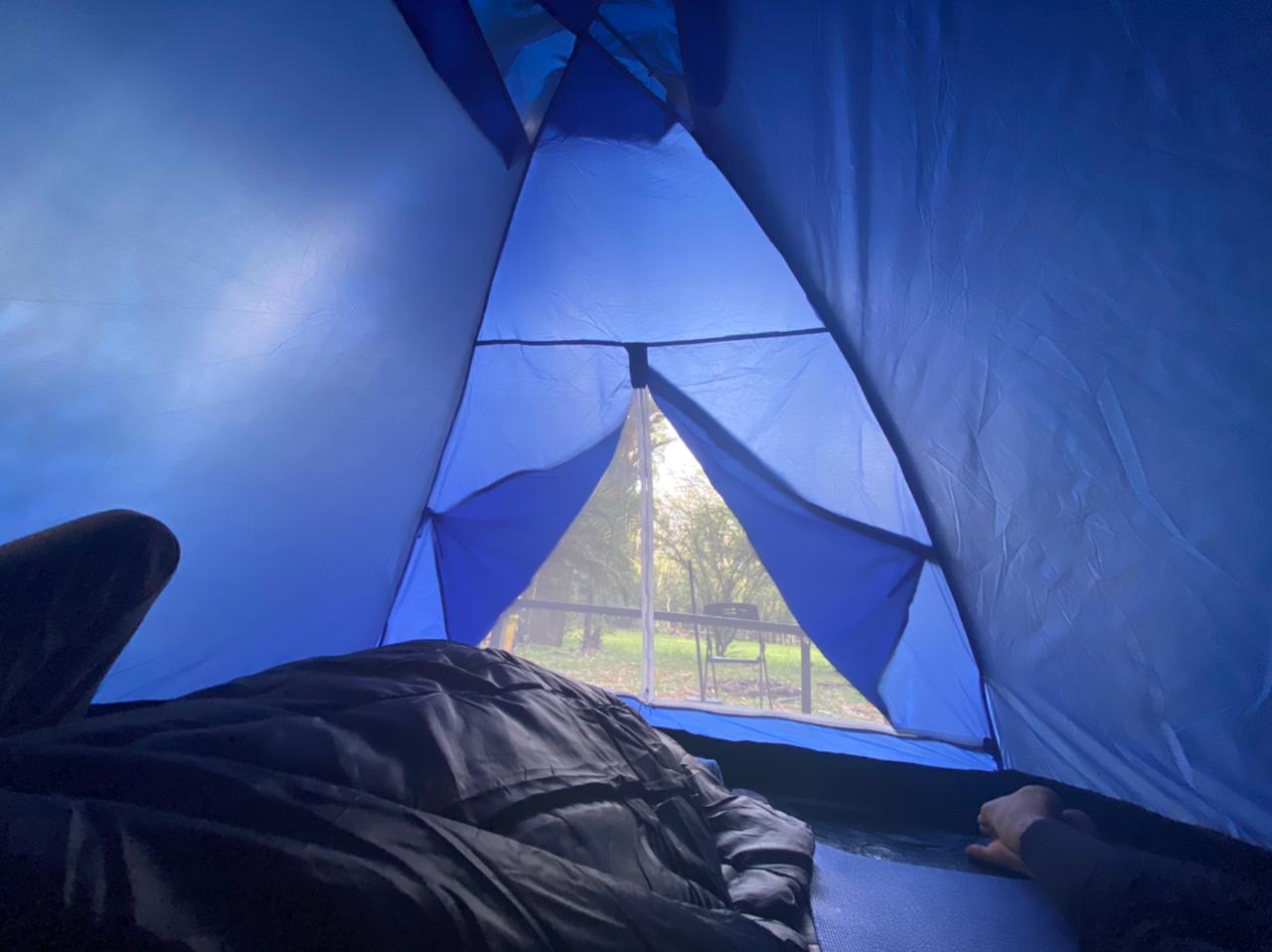 Dentro de una carpa azul, una persona yace mirando hacia afuera. La puerta abierta muestra un parque en un día nublado.