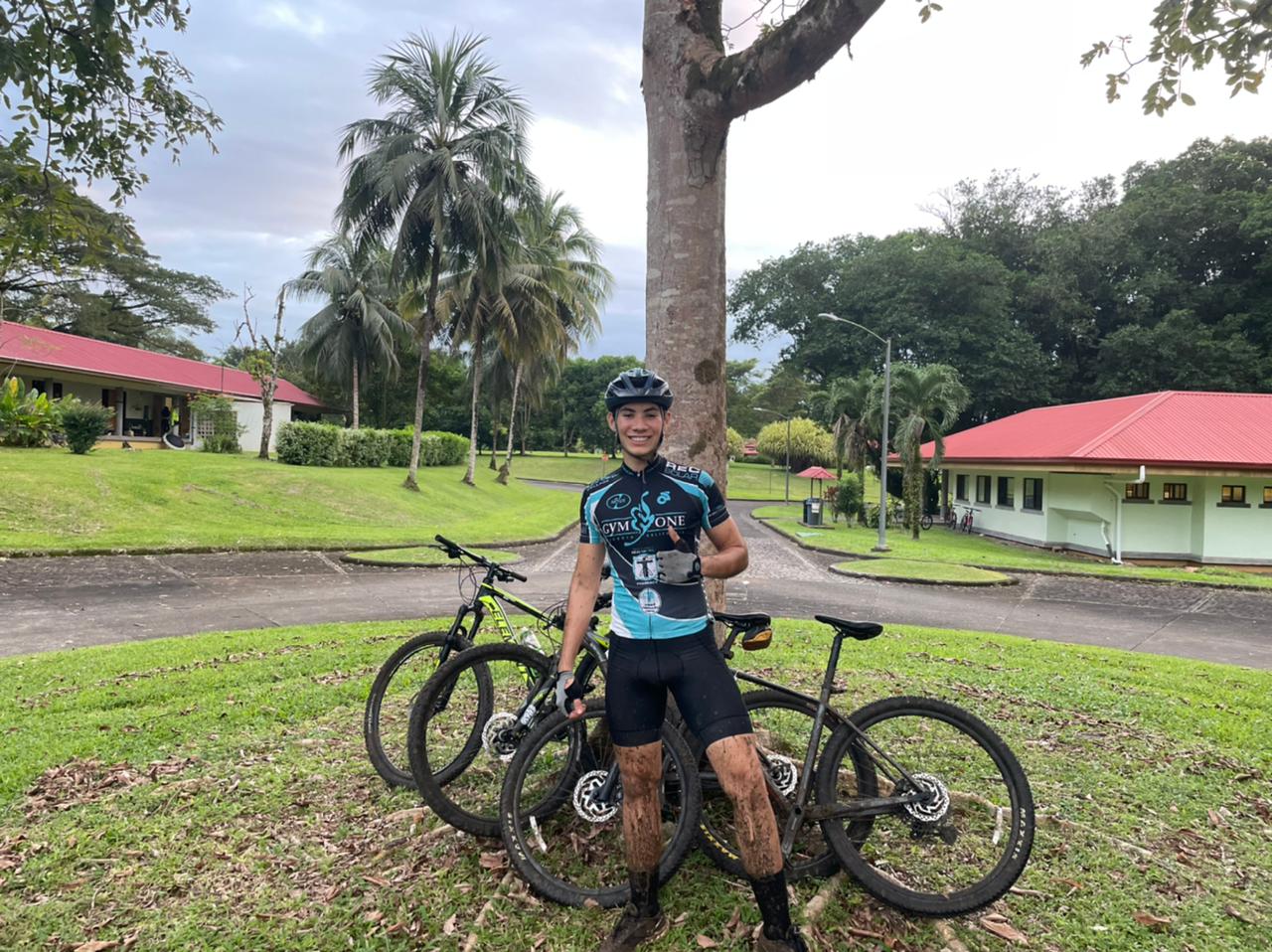 Hombre con dos bicicletas de montaña, sonriendo, delante de edificios y un árbol.