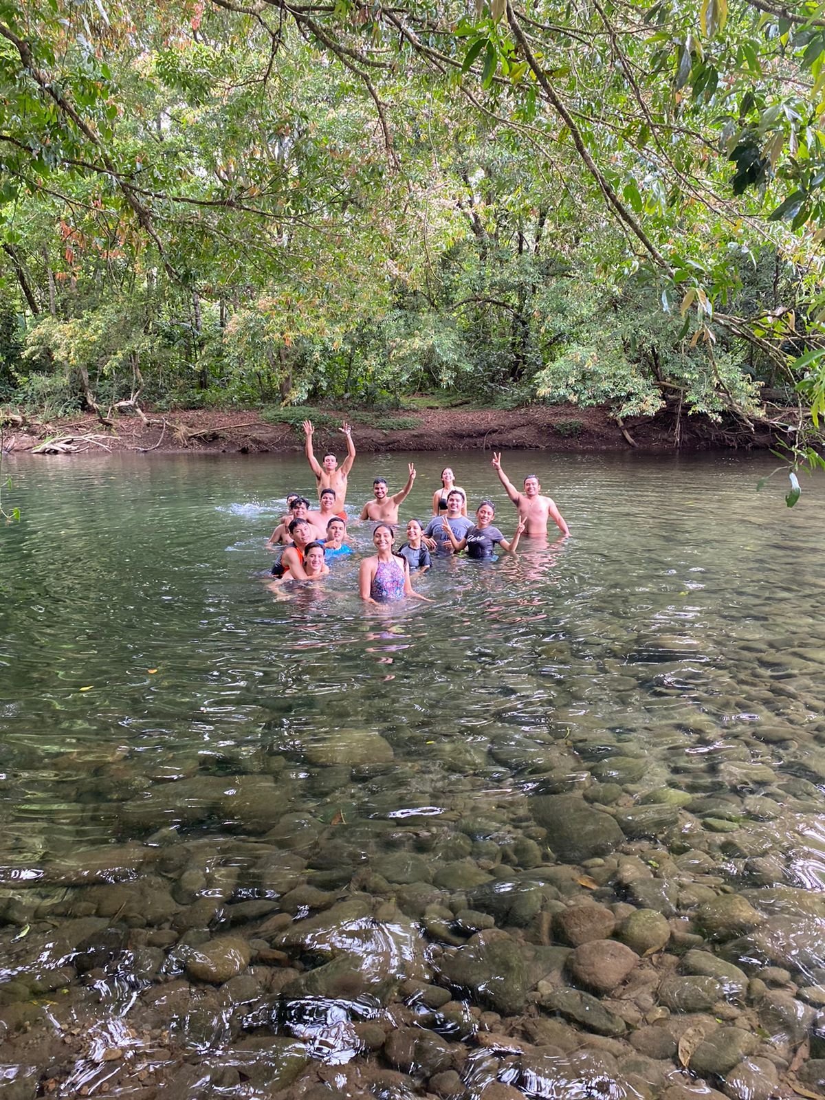 Grupo de personas nadando en un río cristalino, con los brazos en alto. Rodeado de árboles y rocas, en un día soleado.