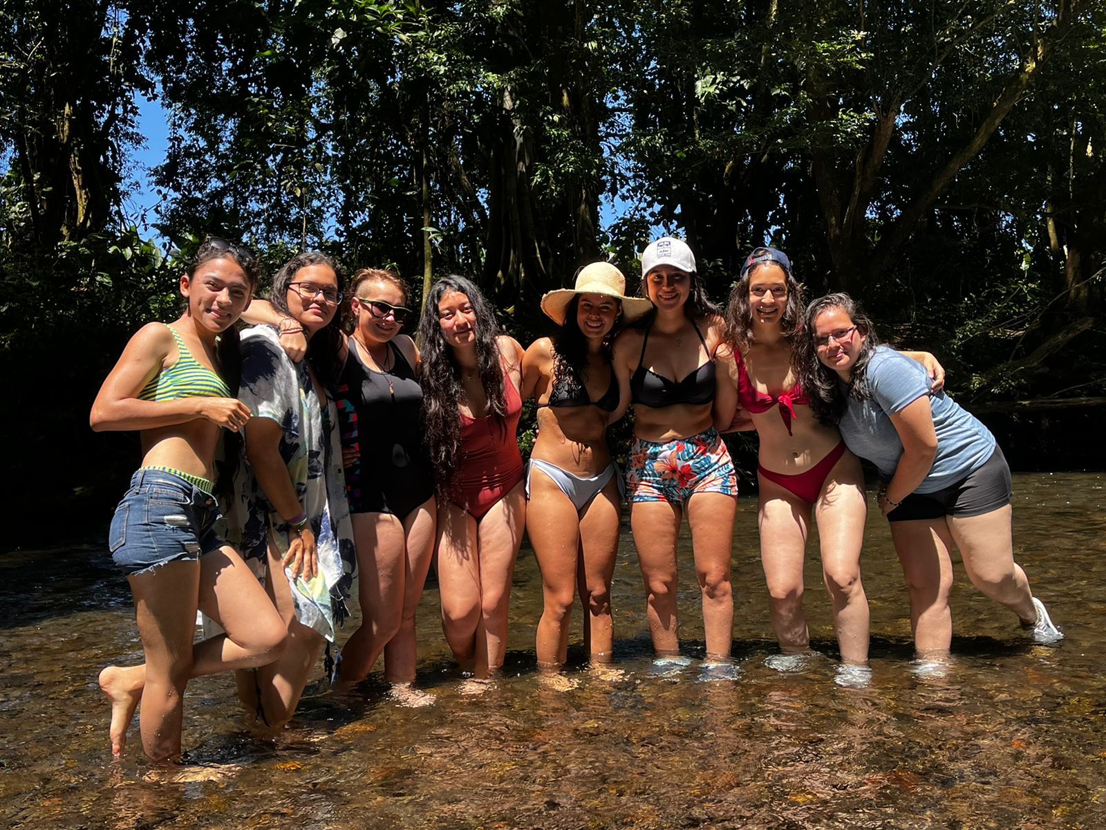 Grupo de mujeres de pie en un río poco profundo, sonriendo, en un día soleado.