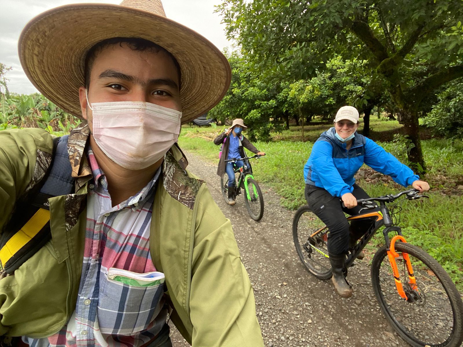 Una persona con sombrero y máscara se toma una selfie con dos personas en bicicleta por un camino de tierra, con árboles verdes de fondo.