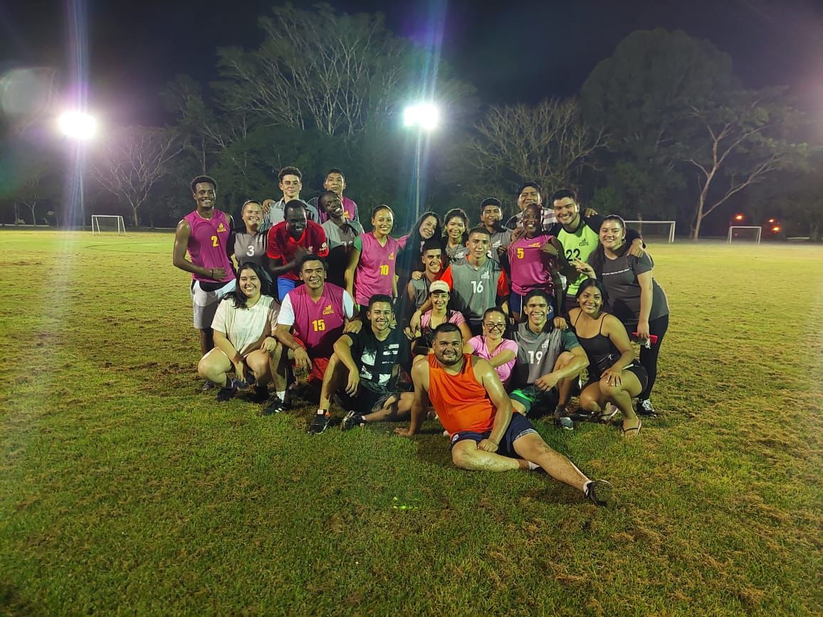 Grupo de personas posando en un campo de hierba por la noche, bajo las luces del estadio. Sonriendo y con ropa deportiva.