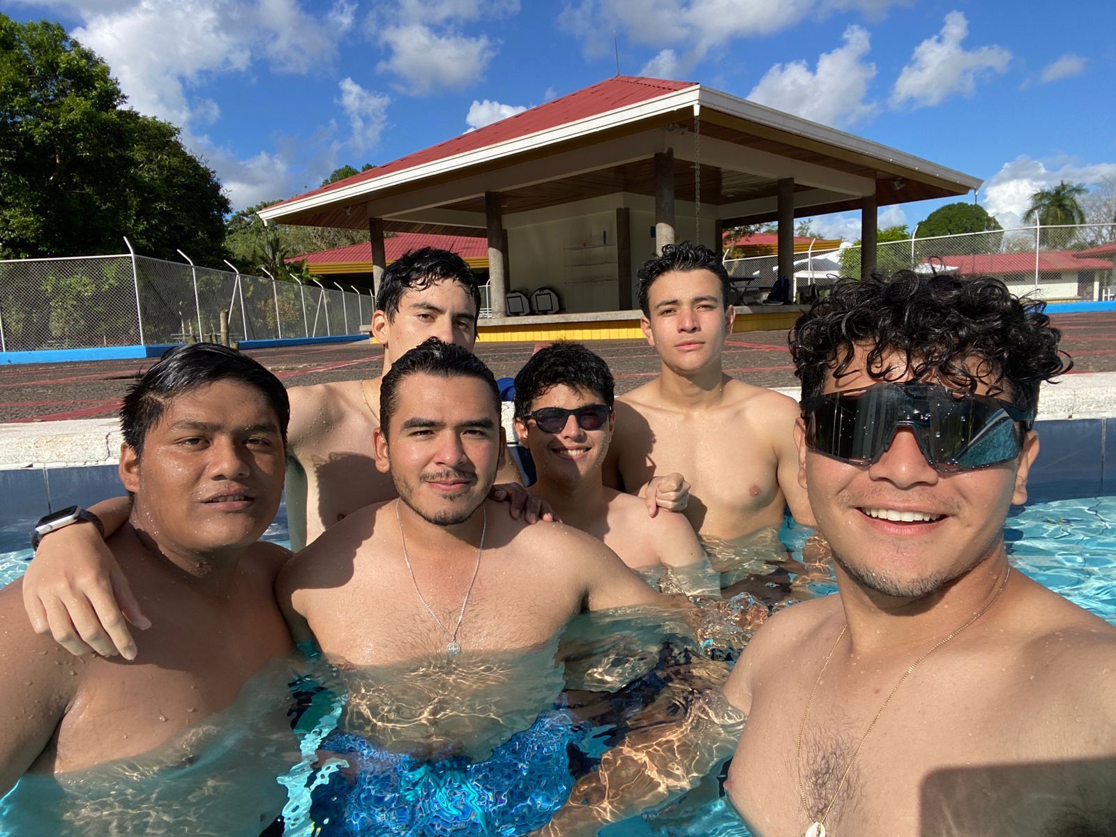 Grupo de seis hombres en una piscina, sonriendo a la cámara. Día soleado, cielo azul.