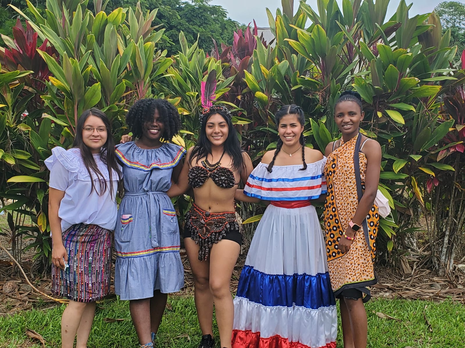 Cinco mujeres con vestidos coloridos posan al aire libre frente a plantas verdes.