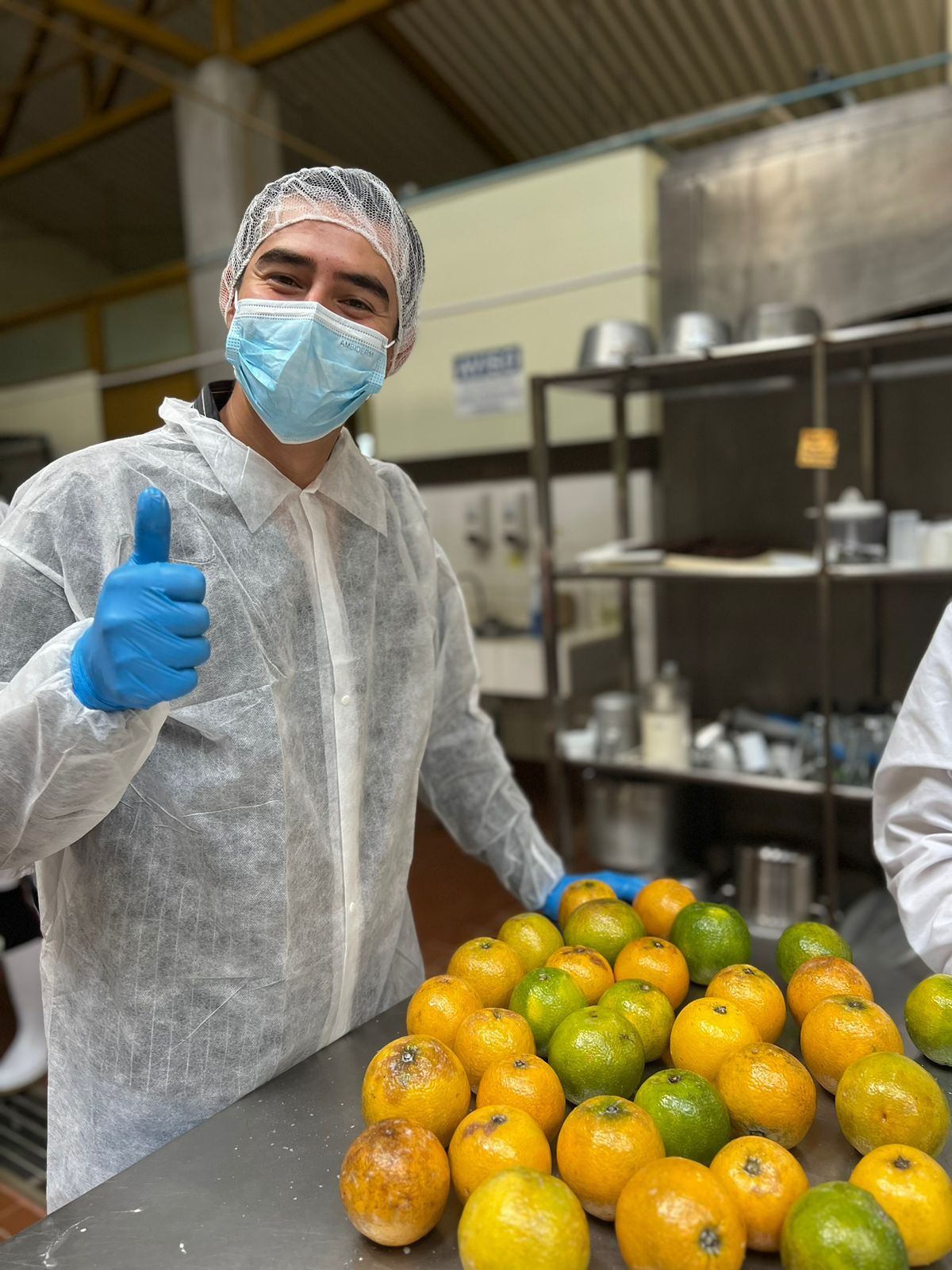 A man wearing a mask and gloves is giving a thumbs up in front of a table full of oranges.