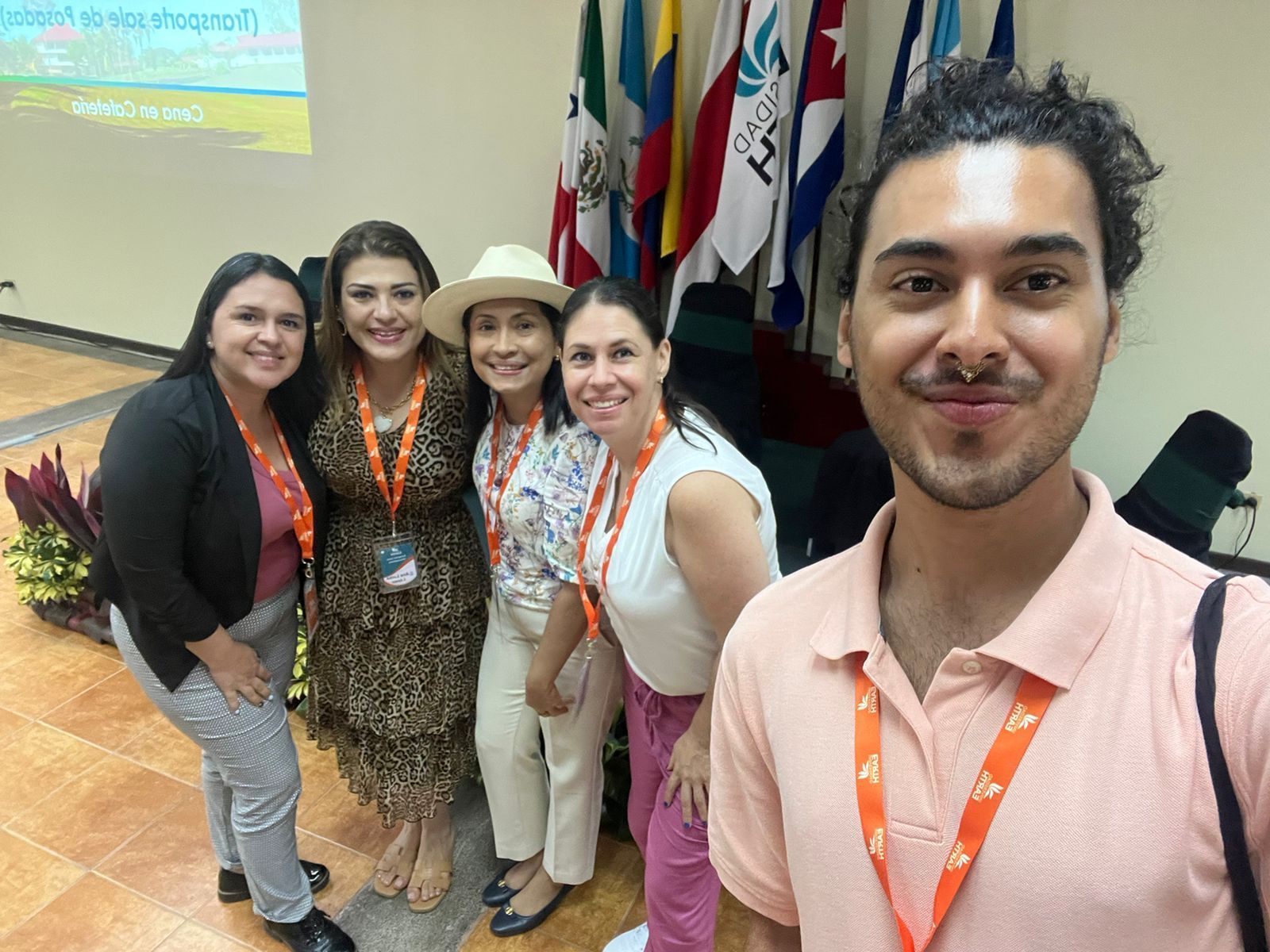 Grupo de seis personas sonrientes posando para un selfie frente a banderas; en el interior.