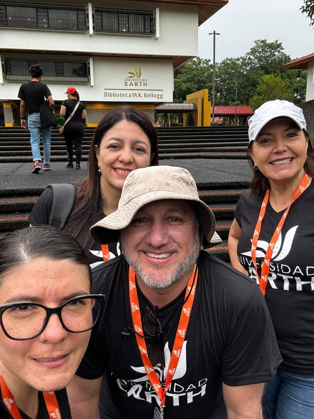 Grupo de cuatro personas sonrientes frente a un edificio; todos llevan el mismo cordón y uno lleva un sombrero.