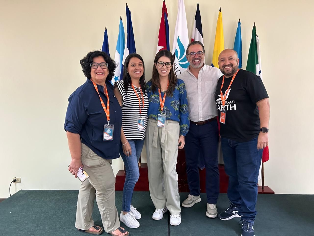 Cinco personas sonrientes, posando frente a banderas. Llevan ropa informal y cordones. Interior.