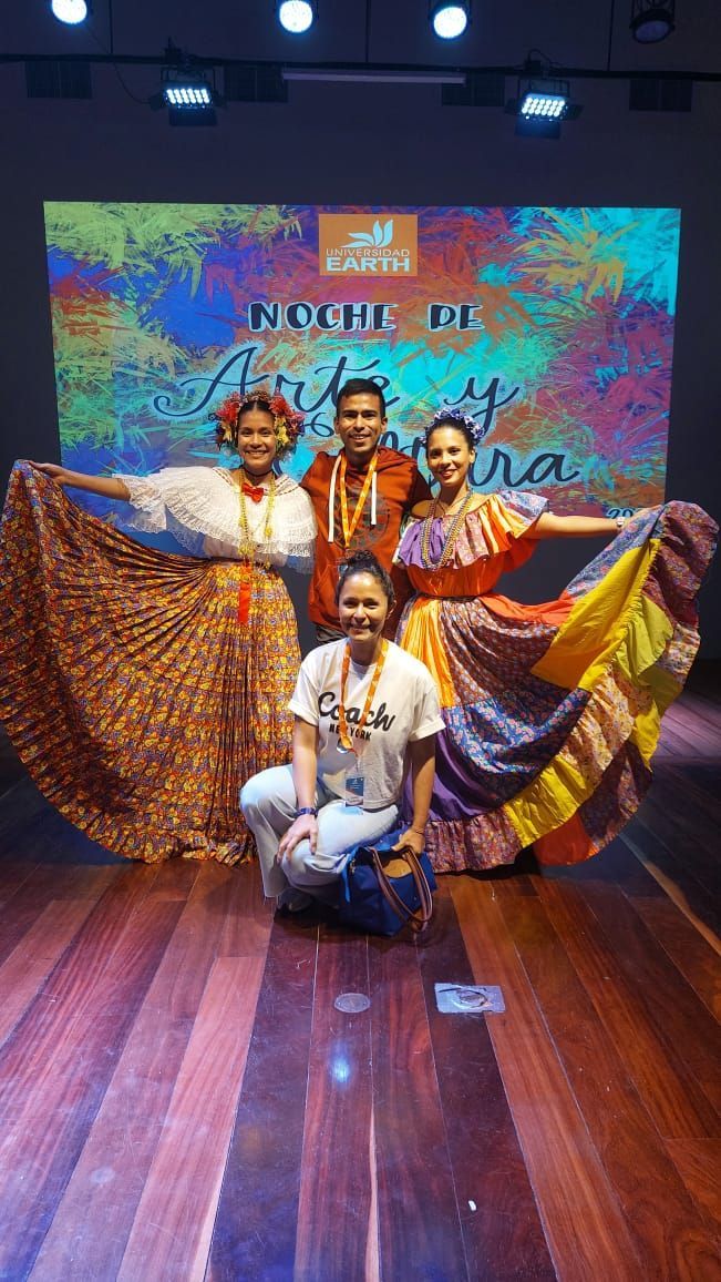 Grupo de personas con atuendos culturales coloridos en el escenario, sonriendo, con un fondo vibrante.