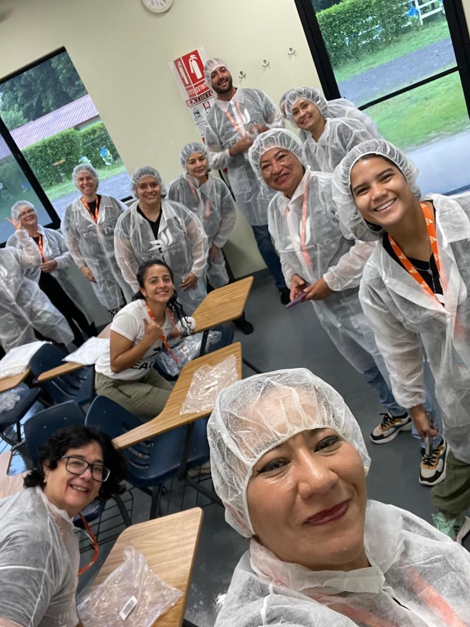 Grupo de personas con redecillas y batas blancas sonriendo en un aula, algunos levantando los pulgares.