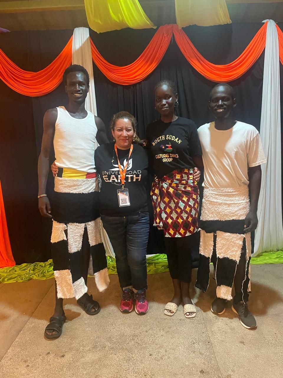 Cuatro personas posan para una foto frente a una tela drapeada. Dos hombres con traje tradicional, una mujer y otra con ropa informal.