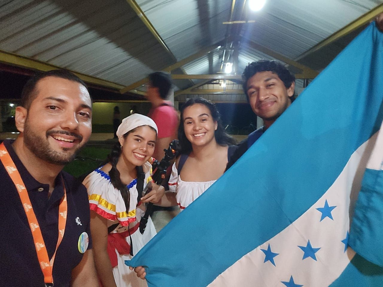 Grupo de personas sosteniendo una bandera hondureña, sonriendo al aire libre por la noche.