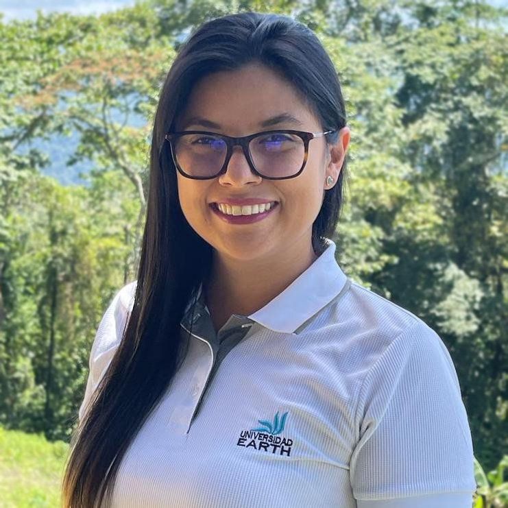 Mujer con gafas, sonriendo con un polo blanco, al aire libre con follaje verde, logotipo en la camisa.