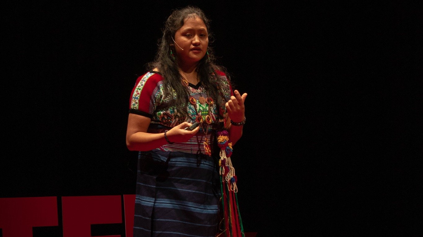 Mujer con atuendo tradicional guatemalteco habla en un escenario TED con fondo negro.