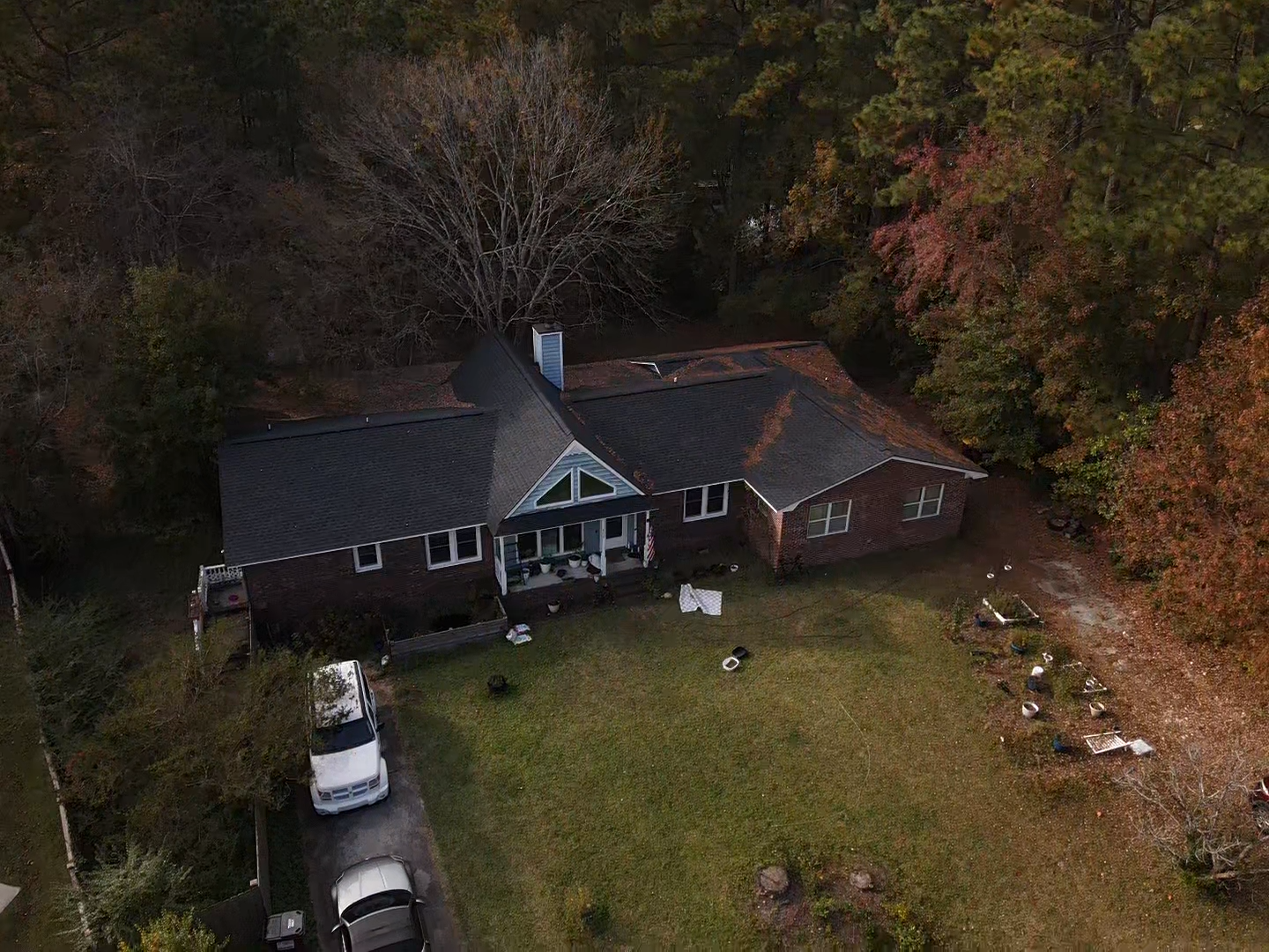 Aerial view of a dark brick house with a black roof, surrounded by trees and a green lawn. Cars parked in the driveway.
